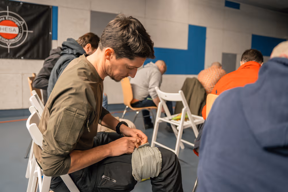 Ein Mann sitzt auf einem Stuhl und wickelt eine Bandage um sein Knie in einem Raum mit mehreren anderen sitzenden Personen.