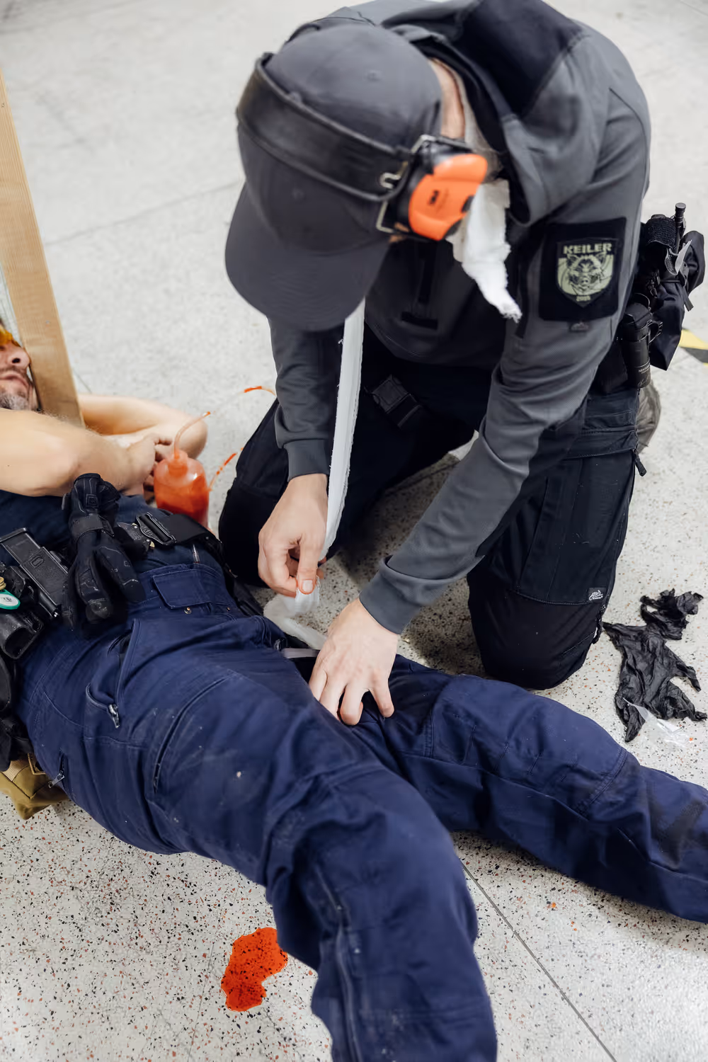 Ein Mann in schwarzer Uniform legt einem am Boden liegenden verletzten Polizisten einen Druckverband an, während sich eine rote Flüssigkeit auf dem Boden verteilt.