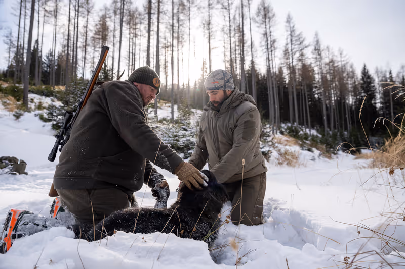 Zwei Männer knien im Schnee in einem Wald und beschäftigen sich mit einem schwarzbraunen Hund, einer der Männer trägt ein Gewehr auf dem Rücken.
