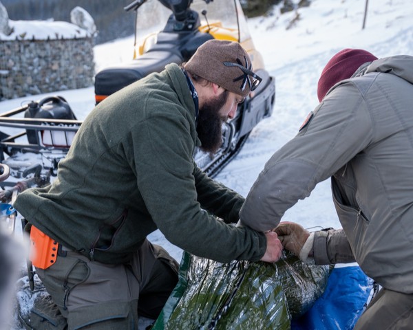 Zwei Männer in Winterkleidung verpacken in der verschneiten Landschaft ein großes grünes Objekt mit Plastikfolie.