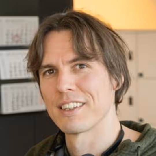 Nahaufnahme eines lächelnden Mannes mit mittellangen braunen Haaren, der eine grüne Kapuzenjacke trägt.