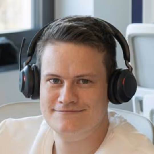 Junger Mann mit kurzen braunen Haaren trägt schwarze Kopfhörer und lächelt leicht.
