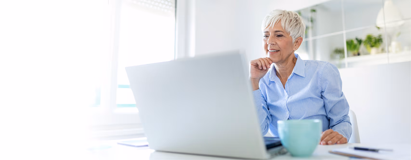Ältere Frau mit kurzem weißen Haar sitzt an einem Tisch und schaut lächelnd auf einen Laptop, mit einer grünen Tasse im Vordergrund.
