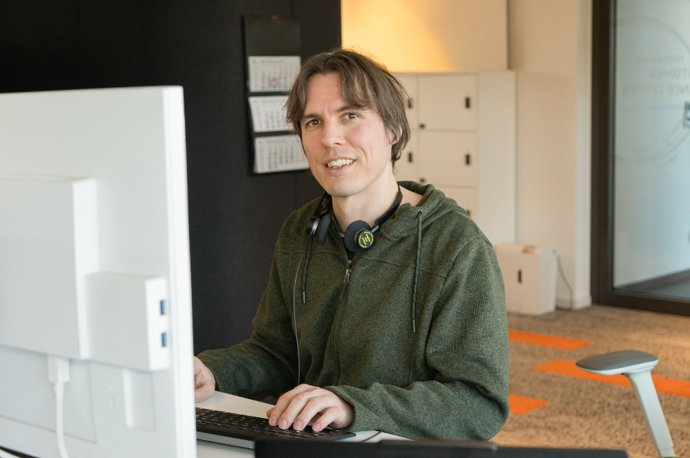 Junger Mann mit Kopfhörern um den Hals sitzt lächelnd am Computerarbeitsplatz in einem modernen Büro.
