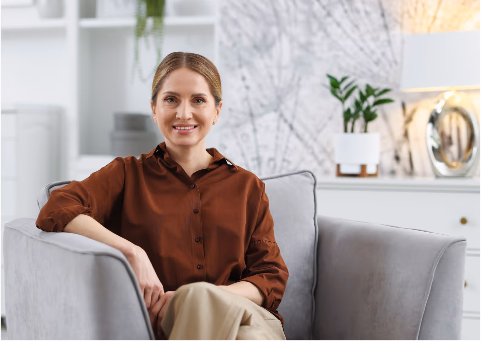 Frau mit braunem Hemd lächelt entspannt auf einem grauen Sessel in modernem Wohnzimmer.