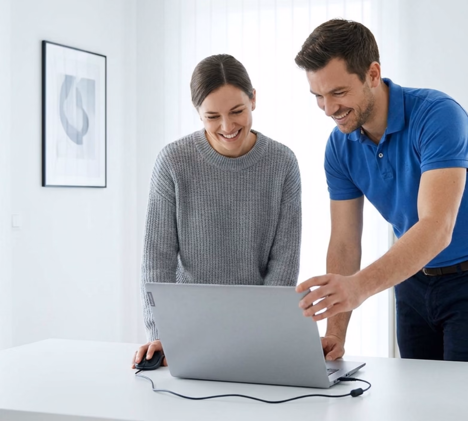 Ein lächelndes Paar steht zusammen vor einem Laptop auf einem weißen Tisch in einem hellen Raum.
