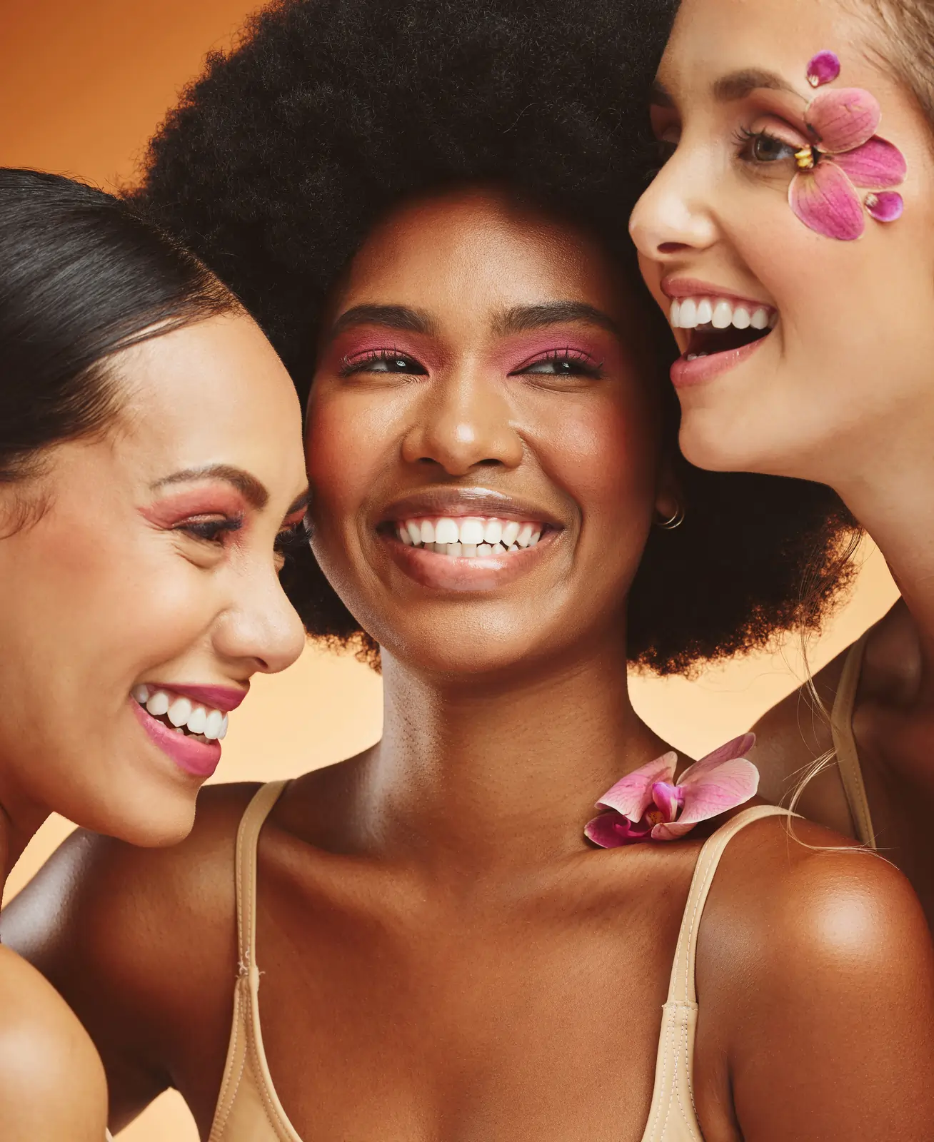 Tre kvinder smiler tæt sammen med pink øjenmakeup og blomster som dekoration.
