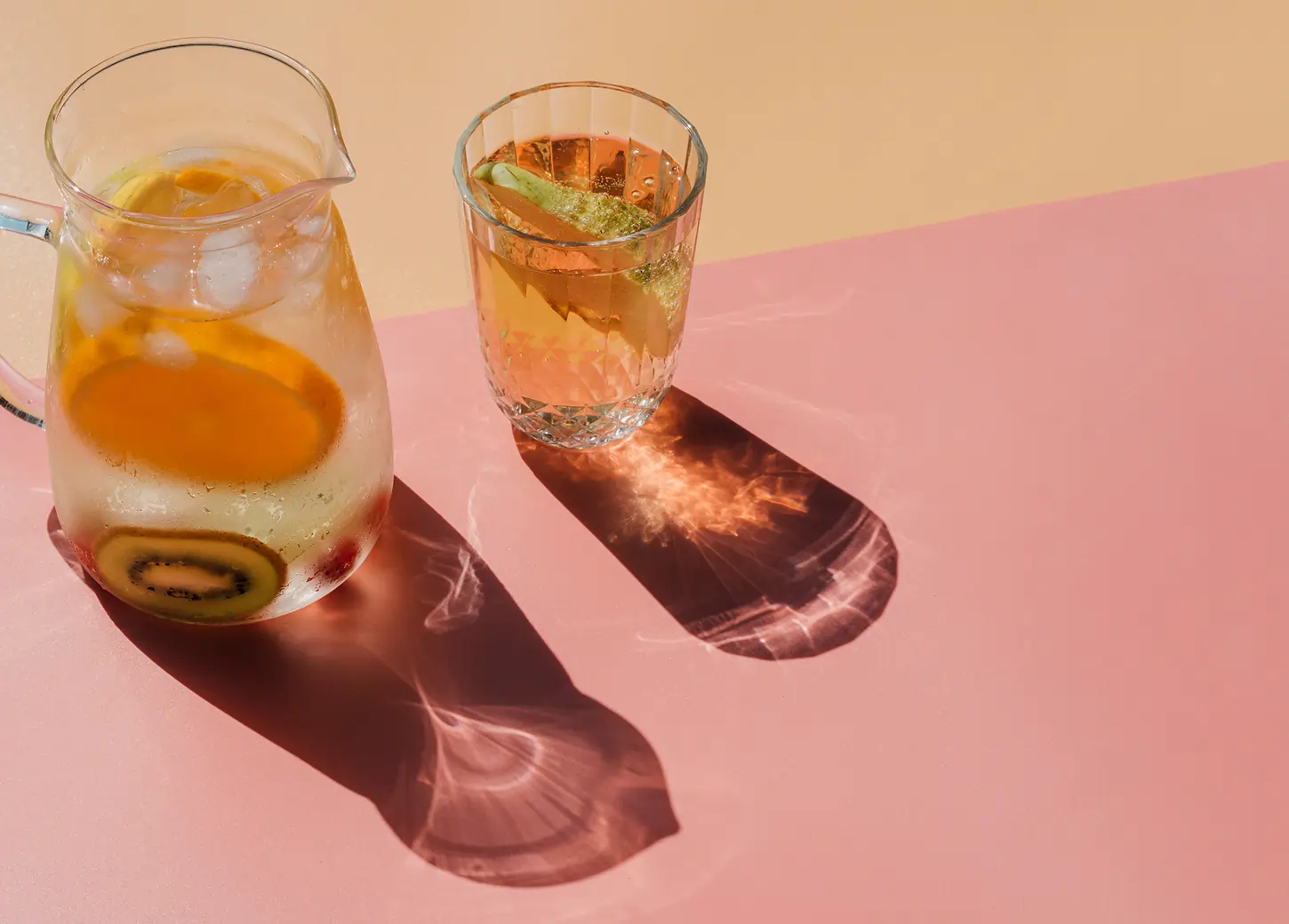 Pitcher and glass of infused water with slices of lemon and kiwi on a pink surface casting shadows.
