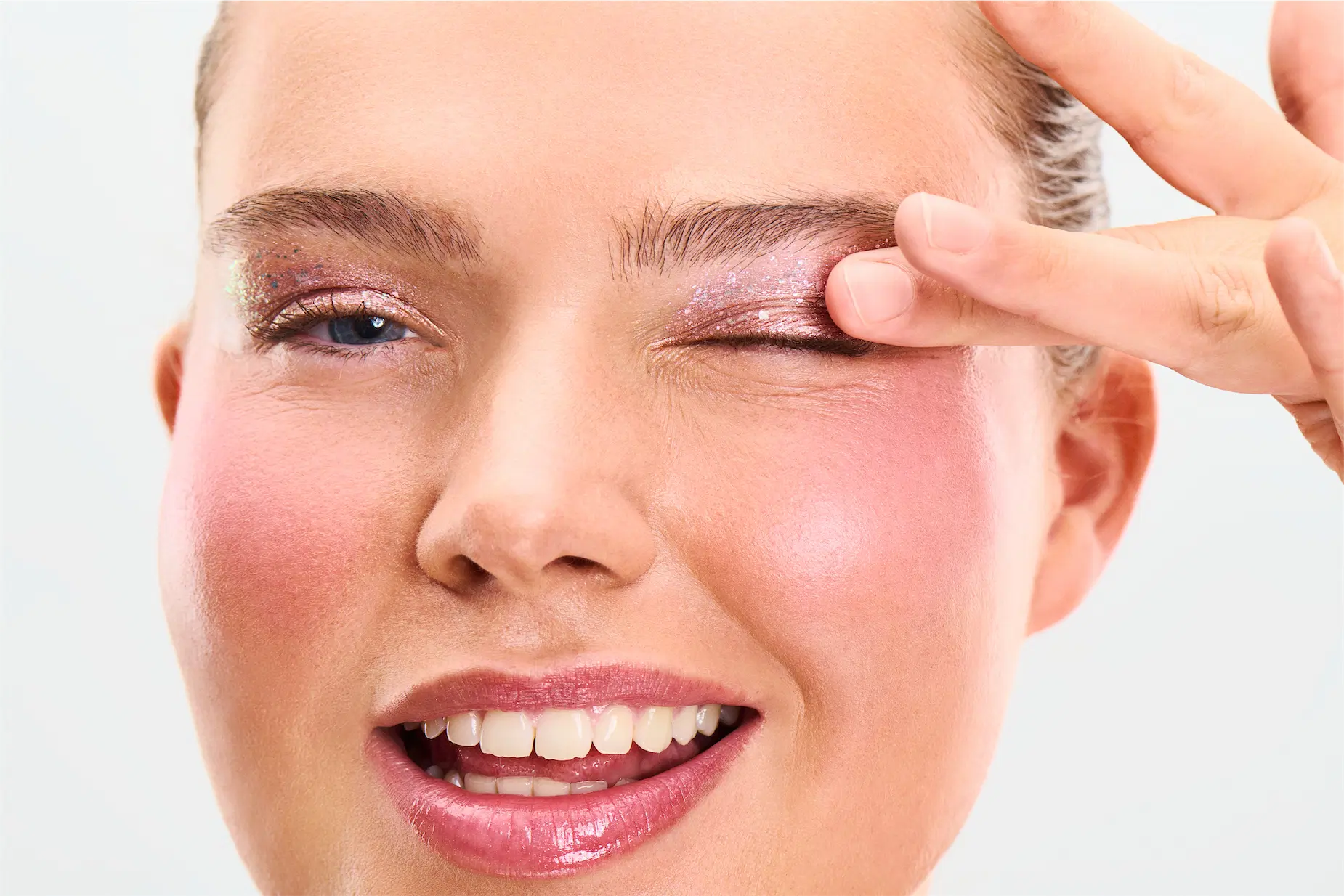 Nærbillede af person med glitrende pink øjenskygge, lyserøde kinder og glossy læber, der lukker det ene øje med fingrene.