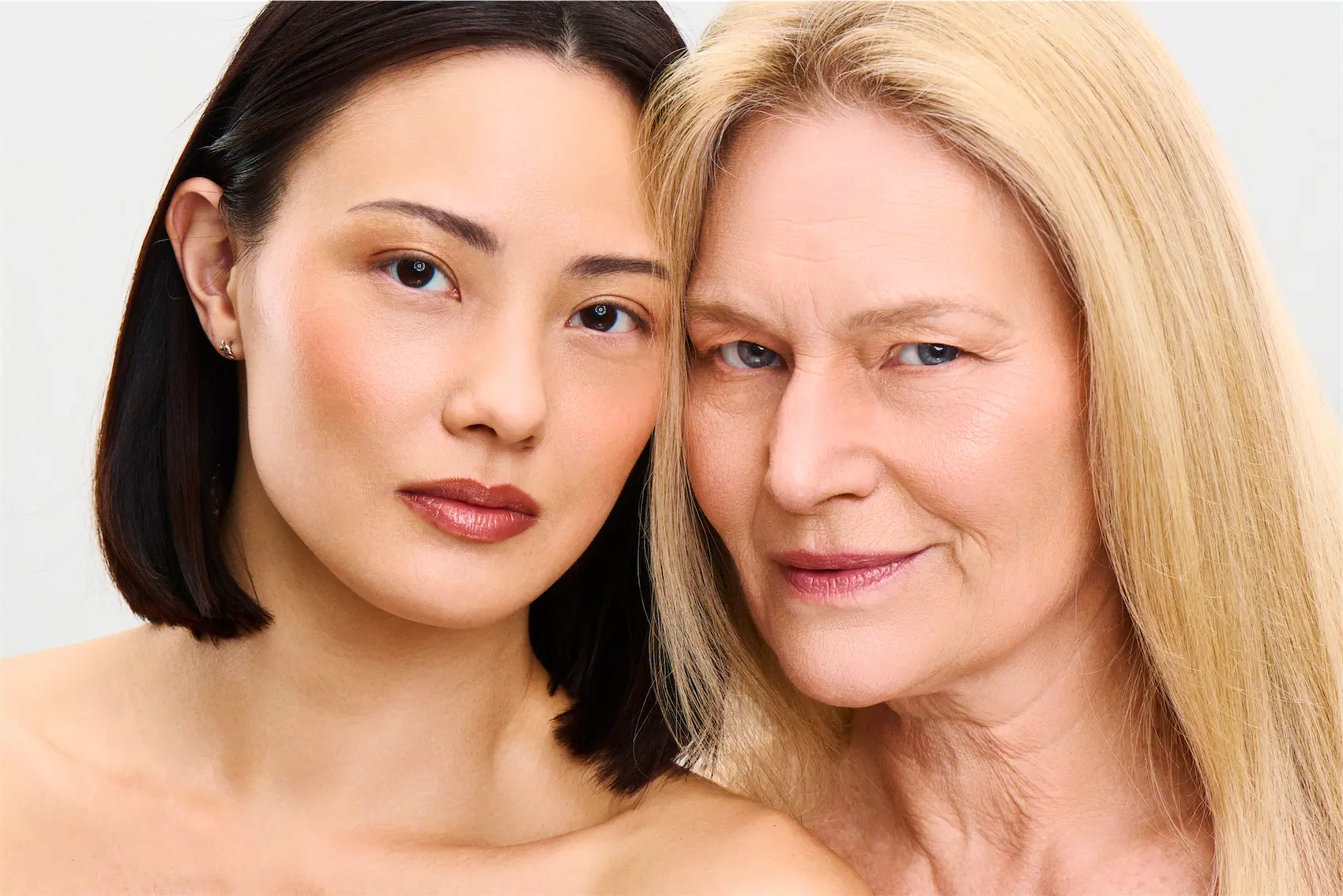 Tre glade kvinder med makeup og blomster, der smiler tæt sammen mod en beige baggrund.