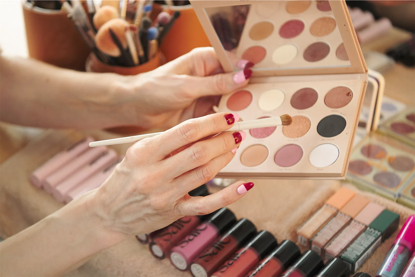 Tattooed arm with swatches of eyeshadow shades near makeup brushes, palettes, lipsticks, perfume, and a round mirror on a white surface.