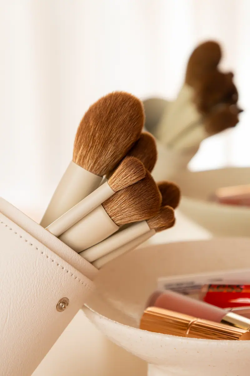 Tre guldmetallic makeup-børster med hvide børstehår placeret på og ved gennemsigtige kvadratiske blokke med to klare kugler foran.