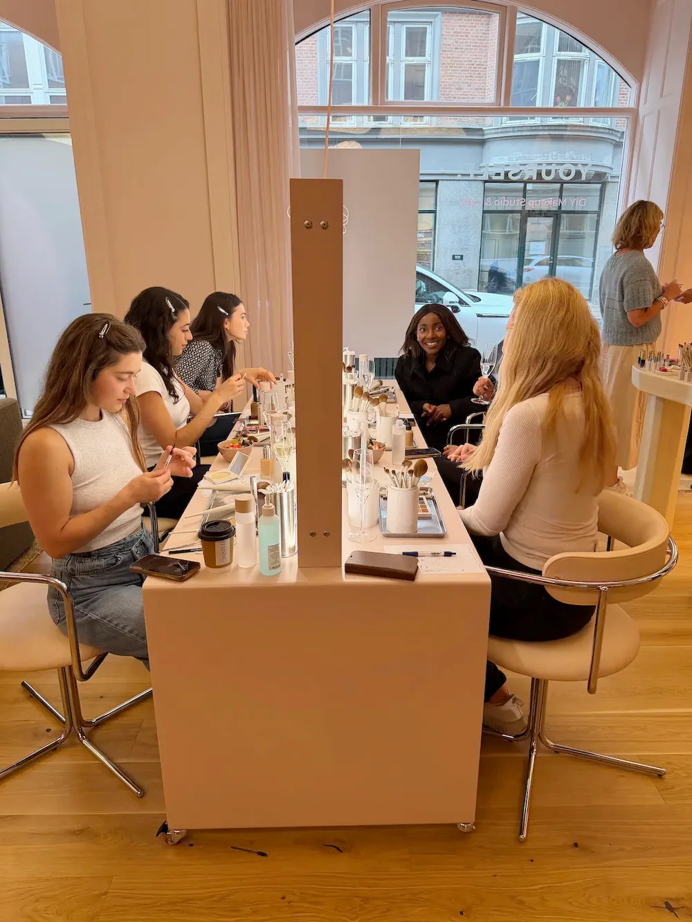 Kvinder sidder ved bord og laver makeup i lyst rum med store vinduer og trægulv.