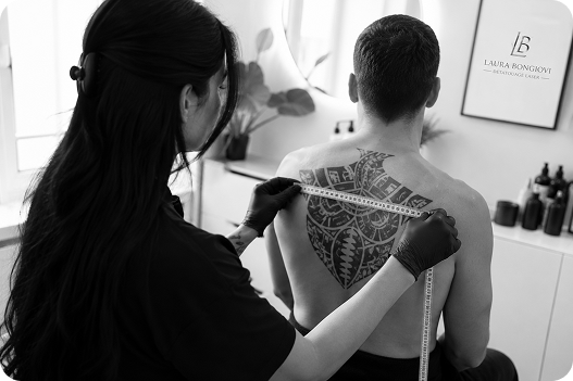 Person with tribal back tattoo being measured across shoulders by a woman wearing gloves in a bright room.