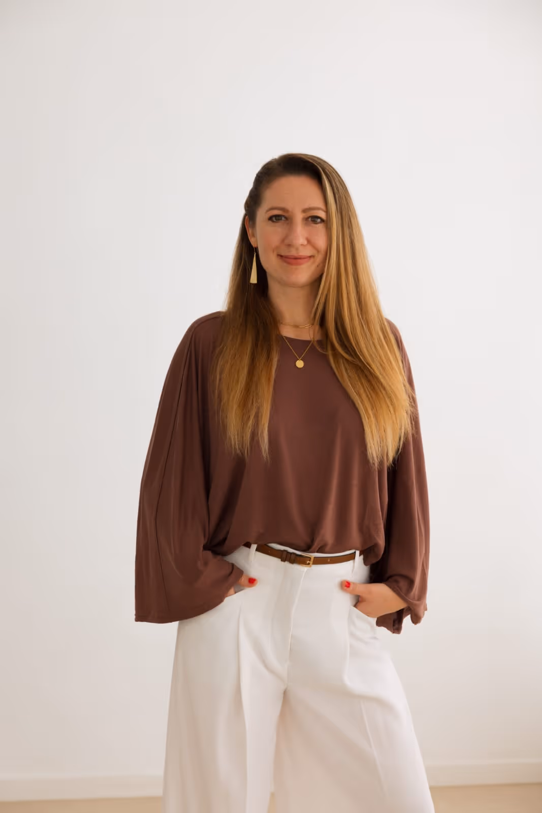 Natalja, smiling, wearing a brown blouse, white pants, and gold jewelry standing against a white background.