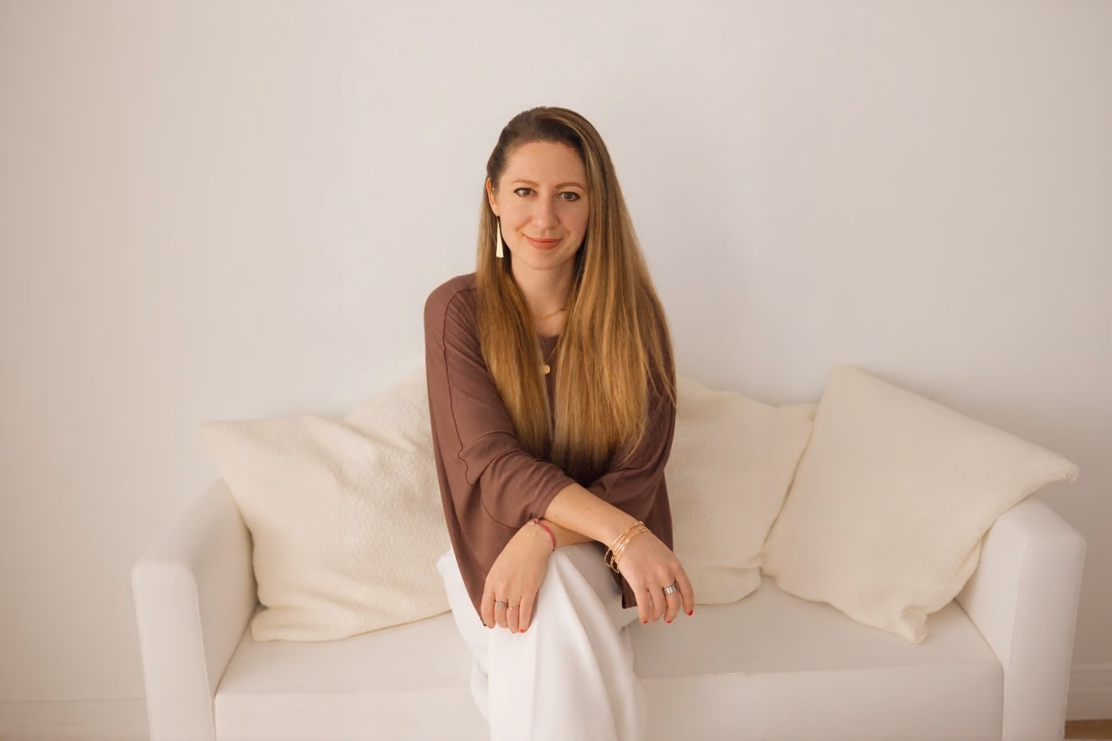 Natalja wearing a brown top and white pants sitting on a white couch with cream pillows, smiling at the camera.
