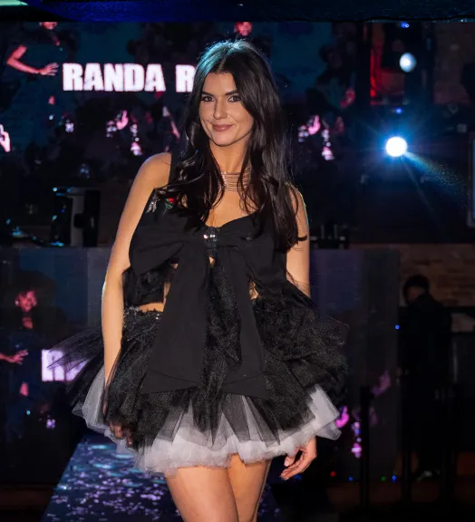 A woman with long dark hair wearing a black and white layered tulle dress with a large black bow on the front, smiling on a runway with illuminated background.