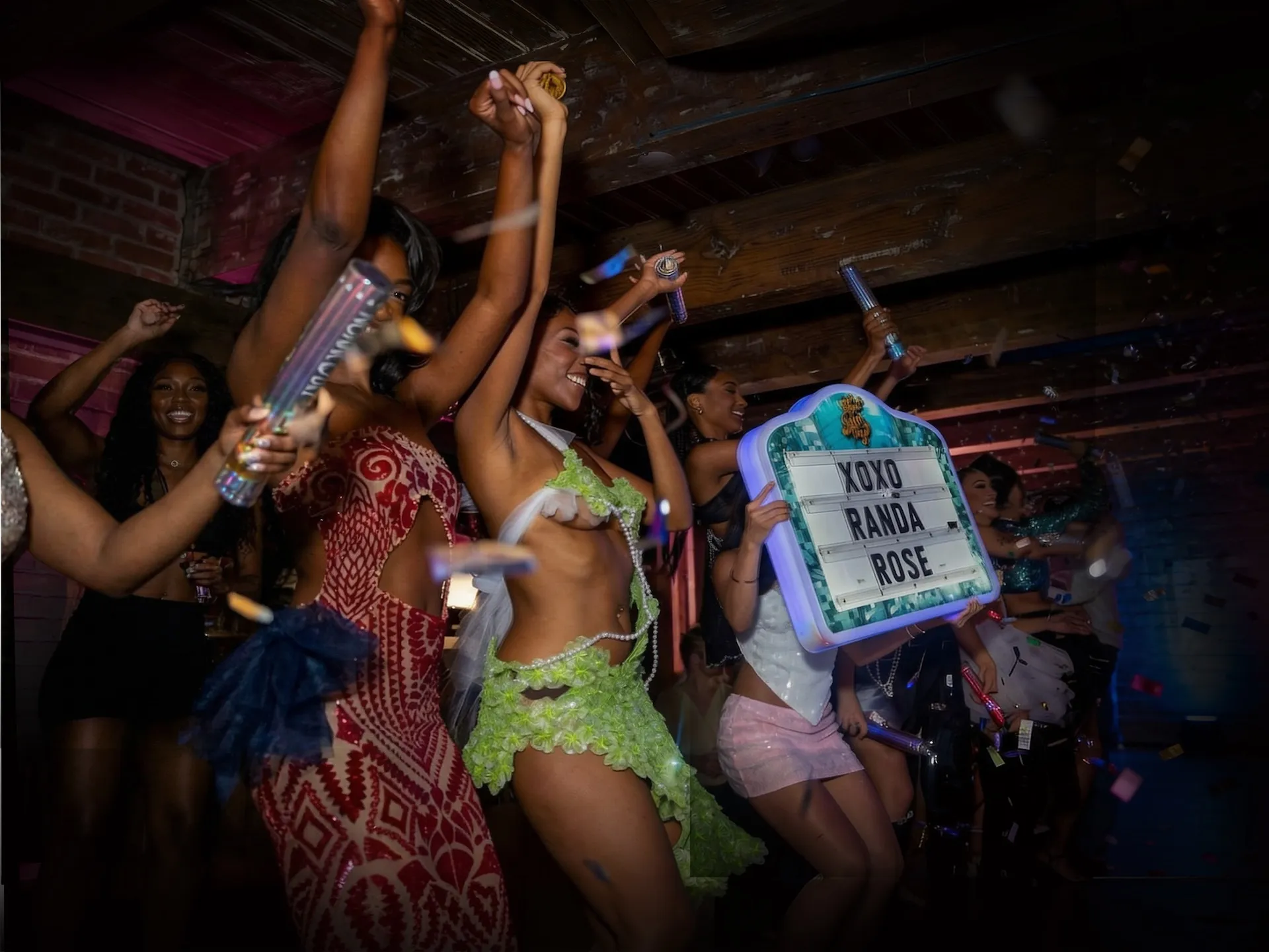 Group of women dancing and celebrating indoors with confetti and party poppers, one holding a sign that reads 'XOXO RANDA ROSE'.