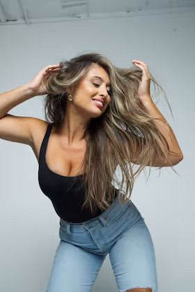 Smiling woman with long hair wearing black tank top and ripped blue jeans posing with hands in her hair.