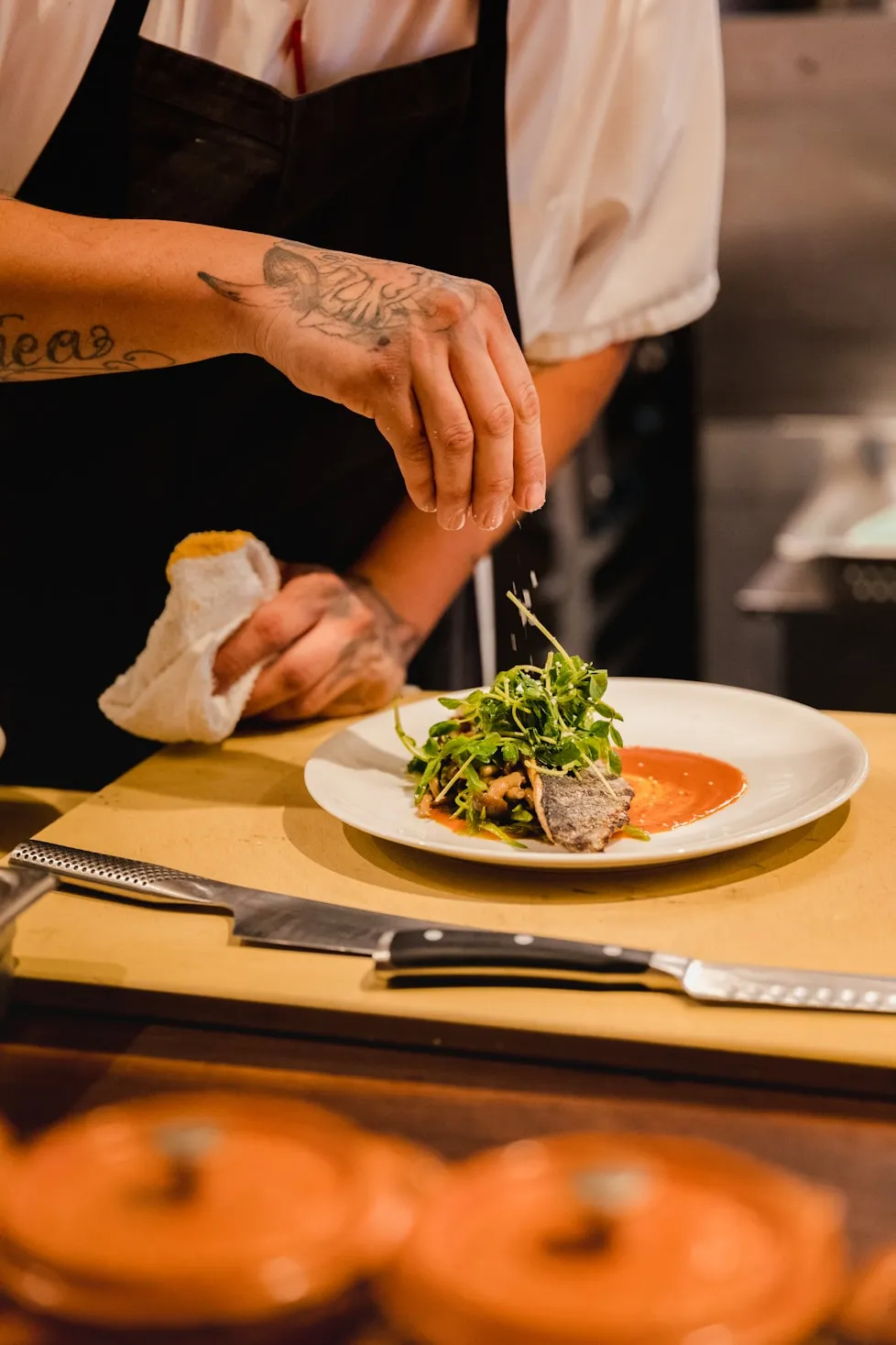 Chef with tattoos sprinkling seasoning over a plated dish with greens and sauce in a kitchen.