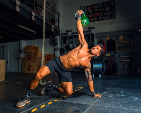 Muscular man in black shorts and gloves performing a side plank while lifting a green kettlebell overhead in a gym.