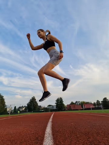 Female athlete in black sports bra and gray shorts jumping mid-air on a red running track with a partly cloudy sky and trees in the background.