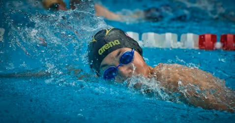 Swimmer wearing a black swim cap and blue goggles swimming in a pool, with water splashing around their face.