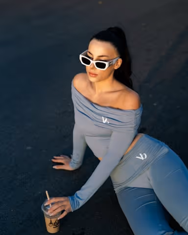 Woman with dark hair in a ponytail wearing white sunglasses and a matching gray off-shoulder long-sleeve top and leggings, sitting and reclining on a dark surface while holding a drink in a plastic cup with a straw.