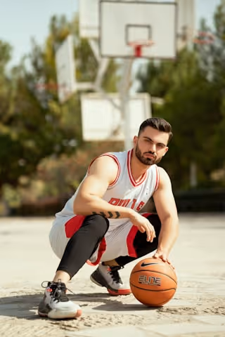 A man in a white and red basketball jersey and black leggings crouches on an outdoor court, resting one hand on an orange basketball with the word 'ELINE', with a basketball hoop and trees in the background.