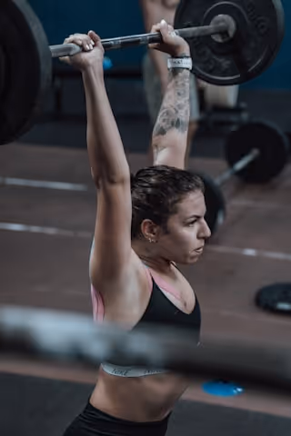 Woman with a tattoo on her left arm lifting a loaded barbell overhead in a gym.