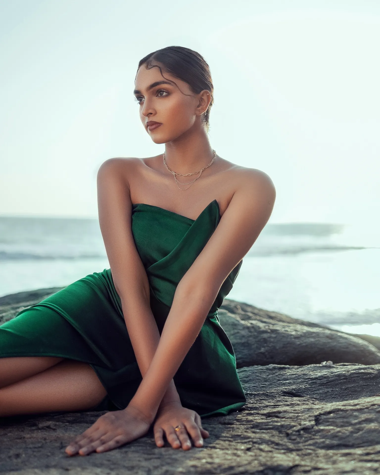 Woman in a green strapless dress sitting on rocks by the ocean at sunset.