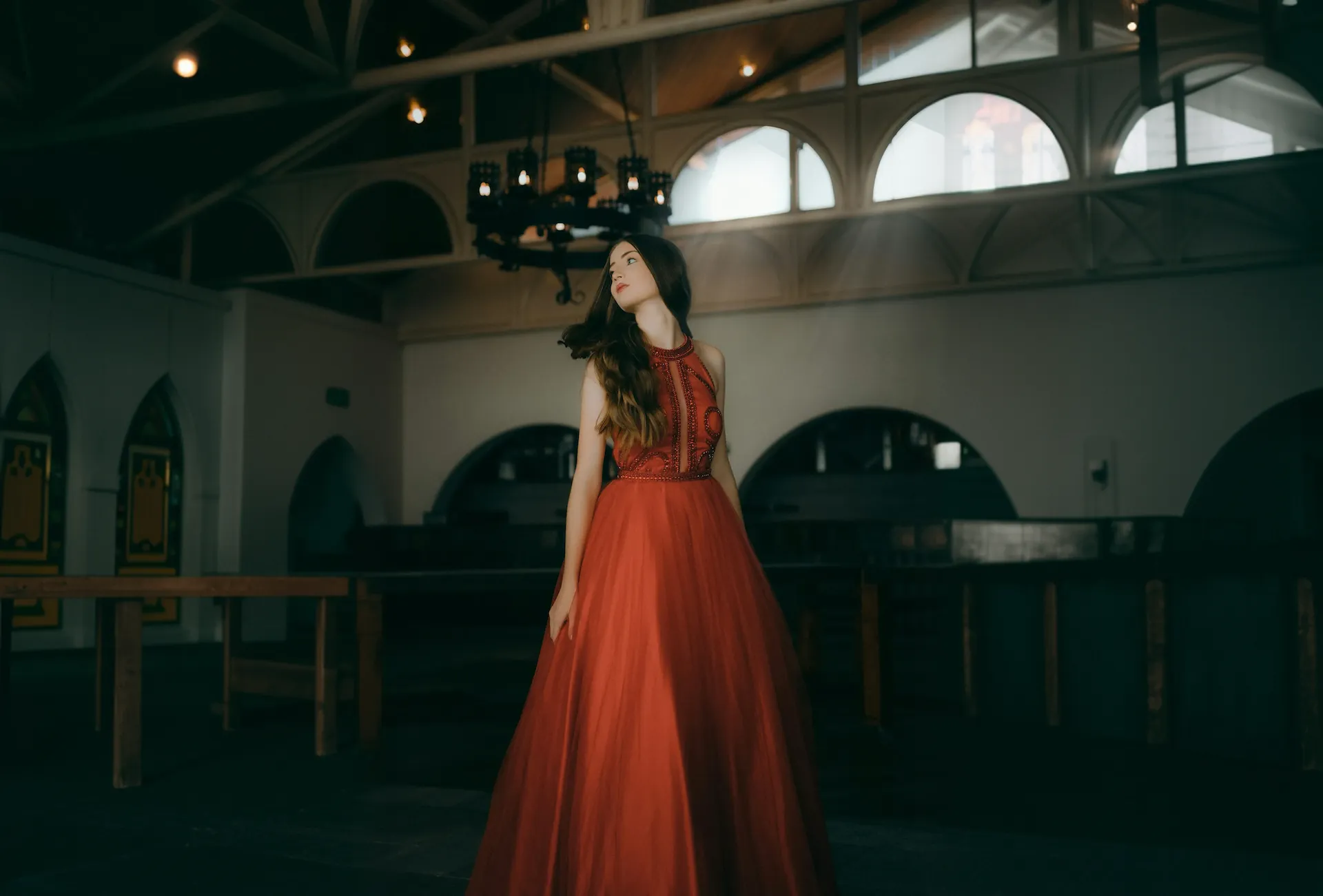 Woman in a flowing red gown standing in a dimly lit room with arched windows and a chandelier.