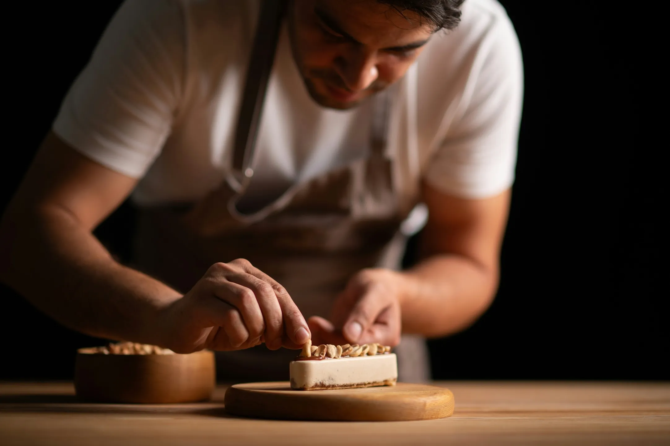 Chef carefully placing nuts on a rectangular dessert on a wooden board in a dark setting.