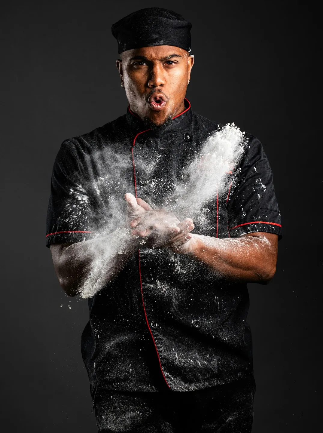 Chef in black uniform clapping hands with flour flying around on a dark background.