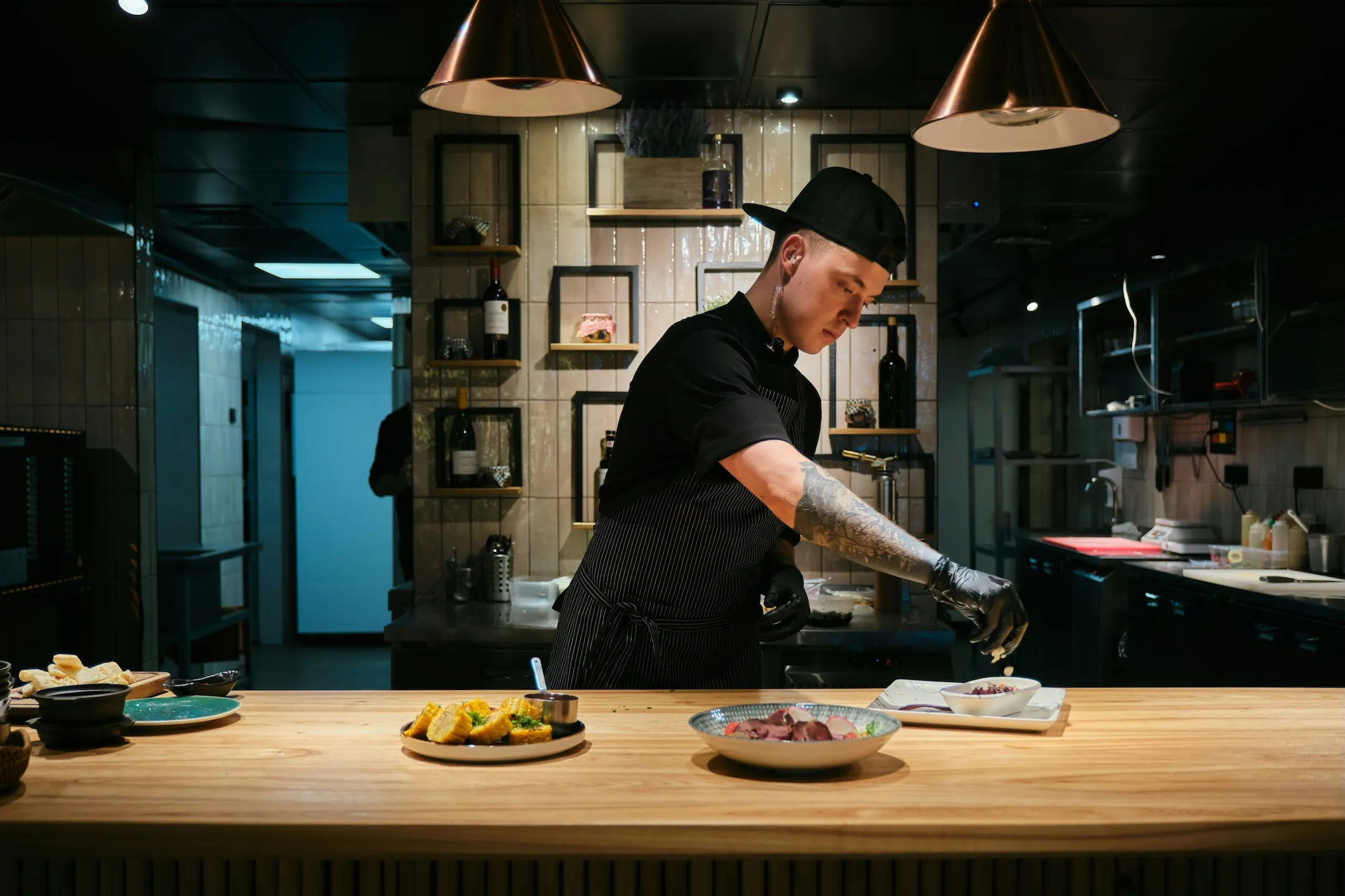 Chef with tattooed arm and black cap garnishing food on a wooden counter in a modern kitchen.