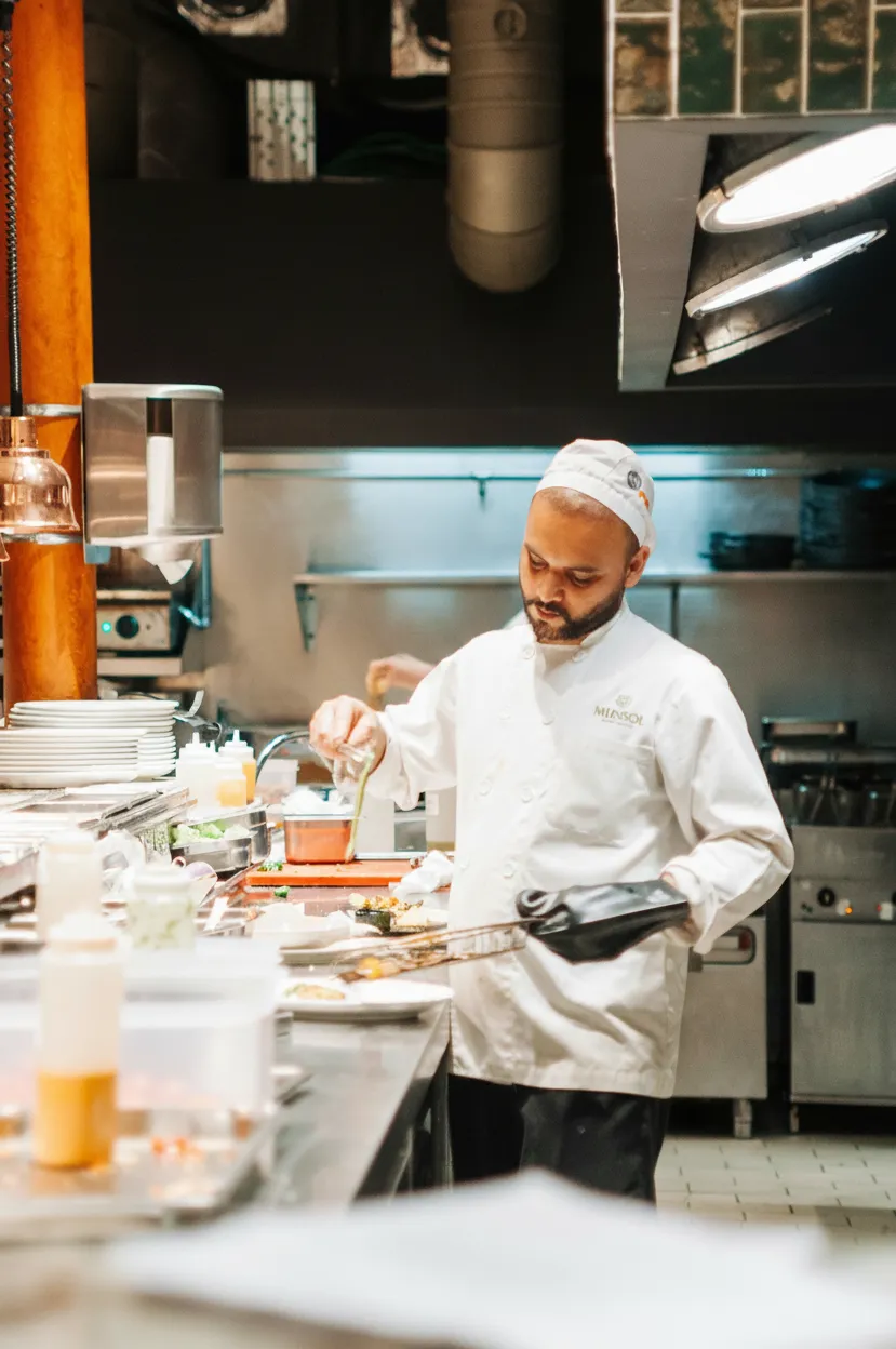 Chef in white uniform plating food with tongs and glove in a professional kitchen.