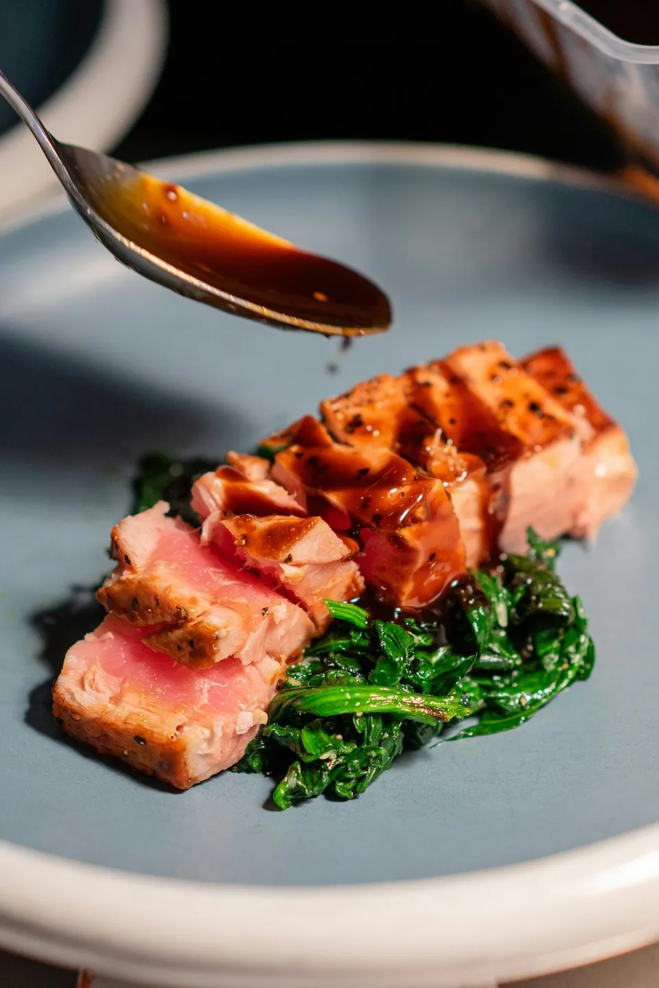 Spoon drizzling sauce over seared rare salmon slices on sautéed spinach on a blue plate.