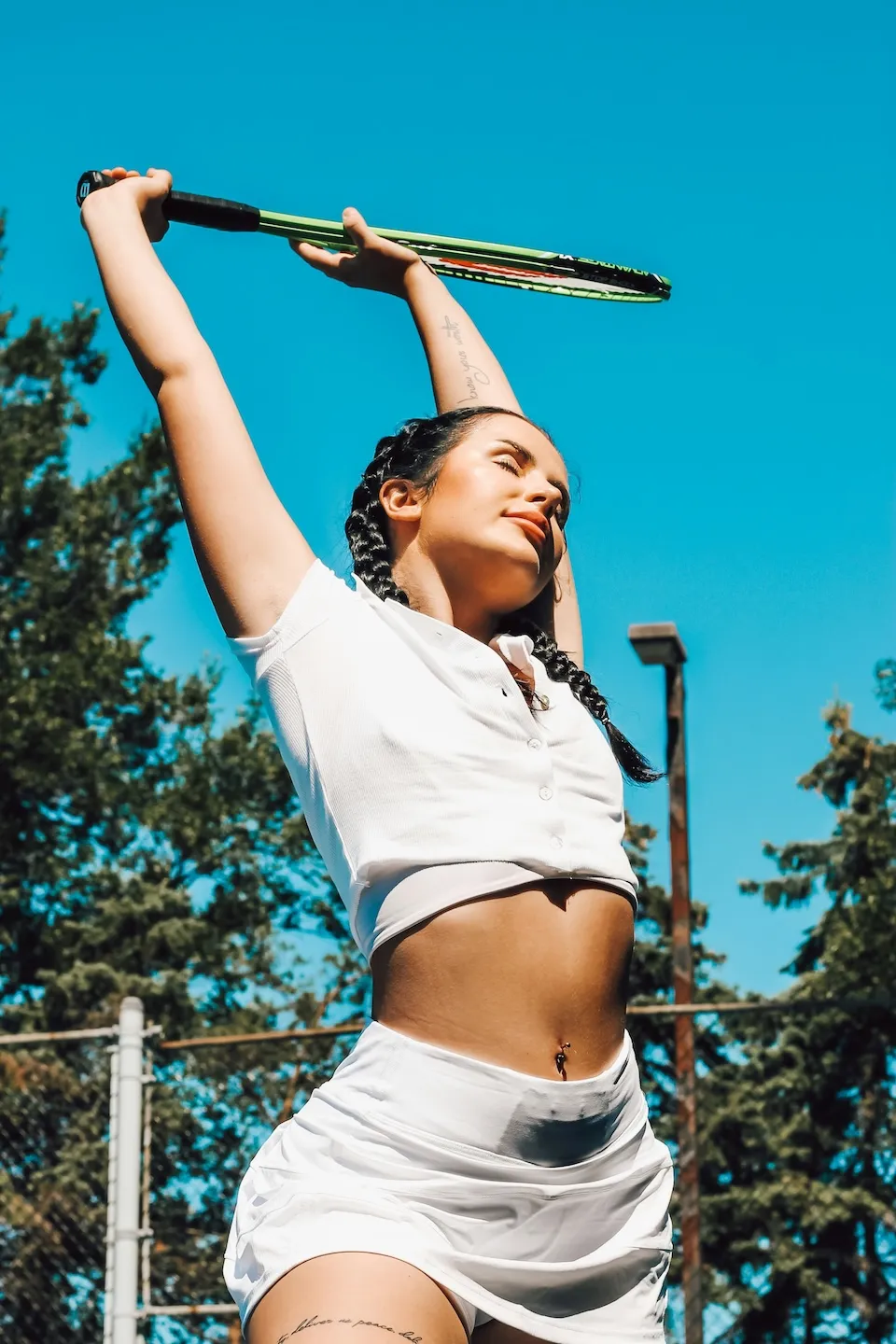 Woman in white athletic outfit stretching with a tennis racket raised overhead outdoors.