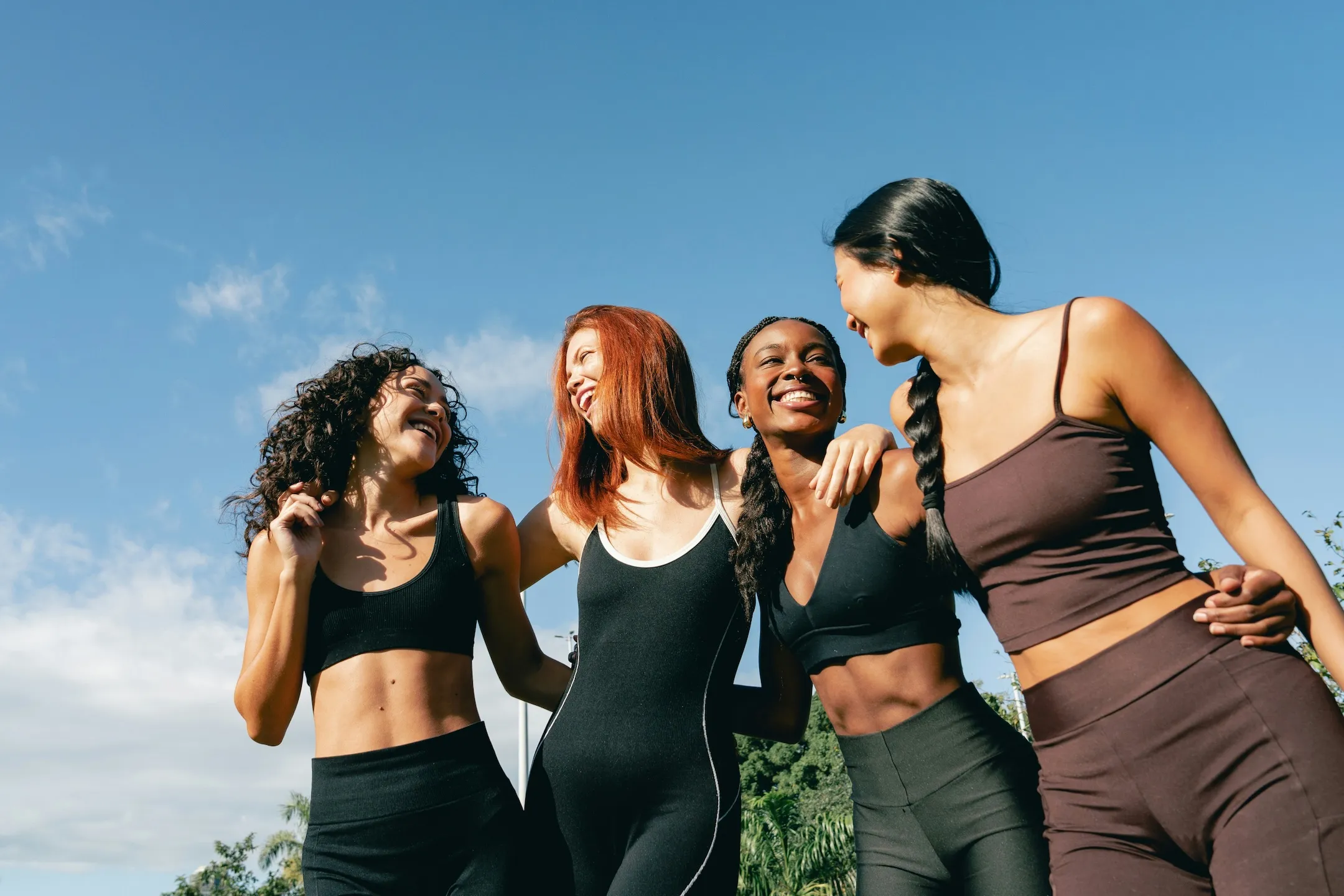 Four diverse women in activewear smiling outdoors under a clear blue sky with arms around each other.