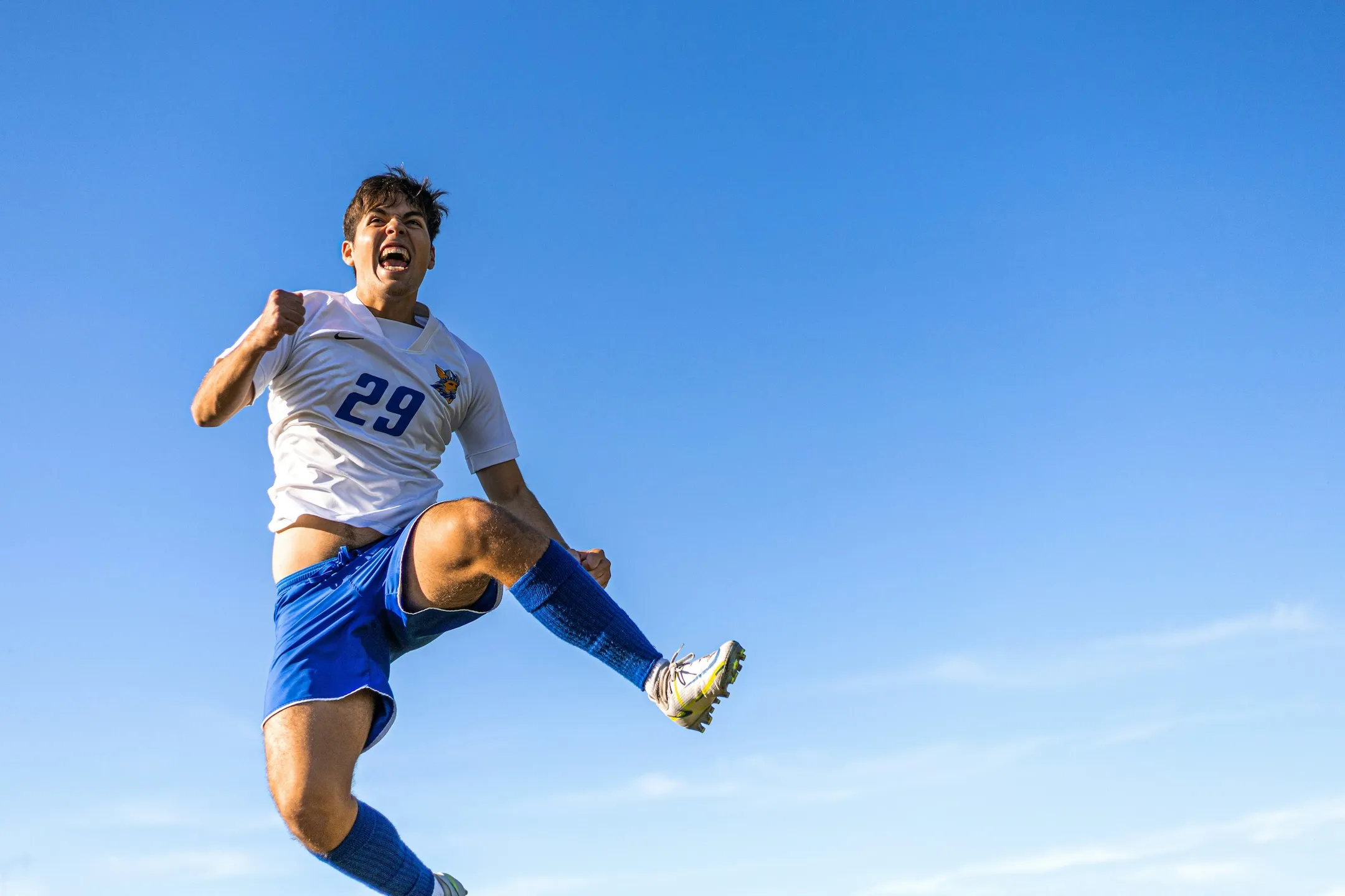 Soccer player in a white and blue uniform jumps with clenched fists under a clear blue sky.