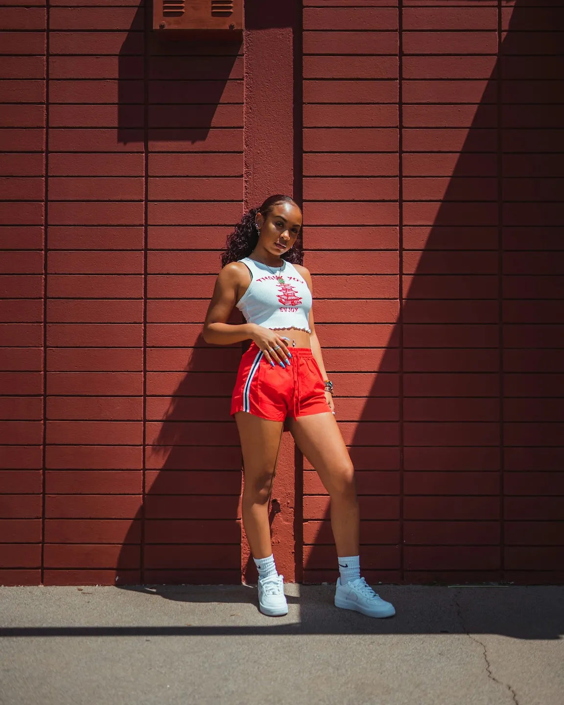 Young woman in red shorts and white crop top standing against a red brick wall in sunlight.