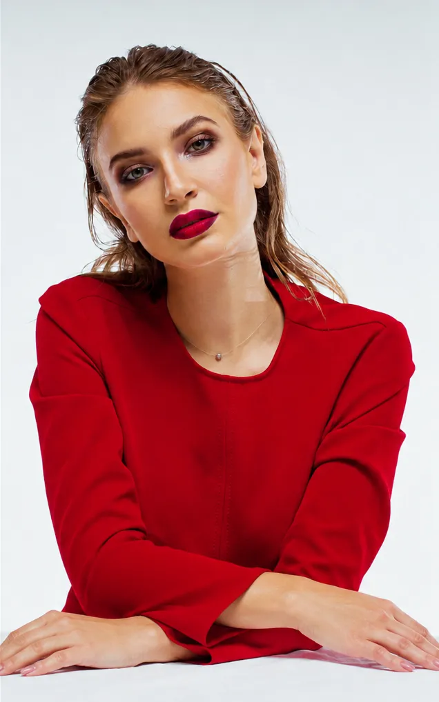 Woman with wet hair and bold makeup wearing a red long-sleeve top, resting her arms on a white surface.