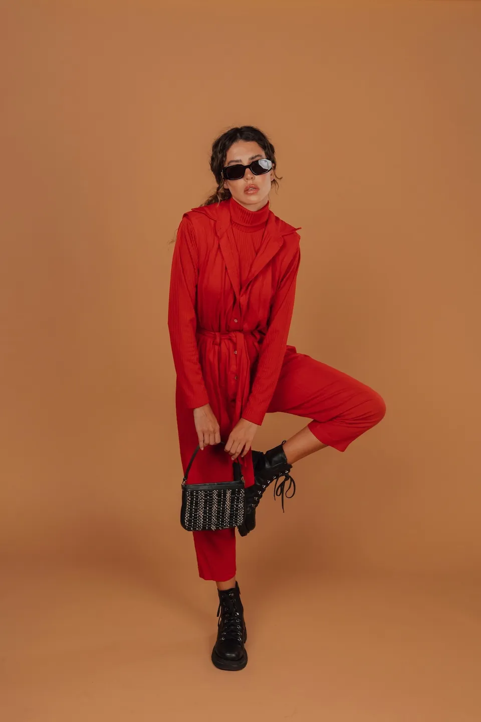 Woman in red outfit and black boots balancing on one leg while holding a small black handbag.