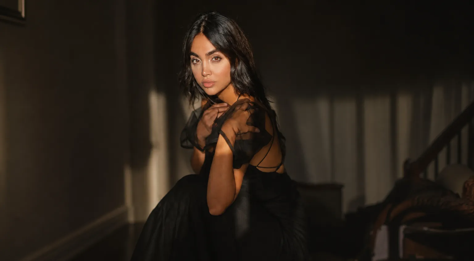Young woman in a black dress sitting inside a dimly lit room with soft sunlight on her face.