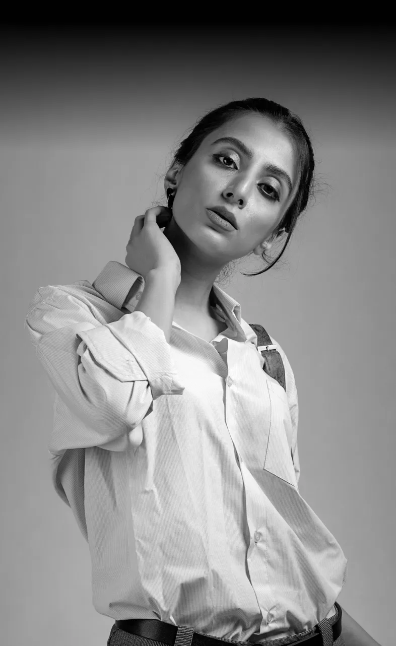 Black and white portrait of a woman with dark hair pulled back, wearing a light button-up shirt with rolled-up sleeves and a belt, looking slightly downward with a calm expression, her right hand touching the side of her neck, against a plain gradient background.