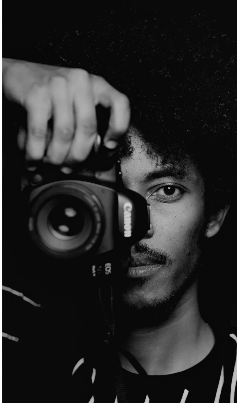 Black and white close-up of a man with curly hair holding a Canon camera close to his face, with one eye visible looking through the camera lens.