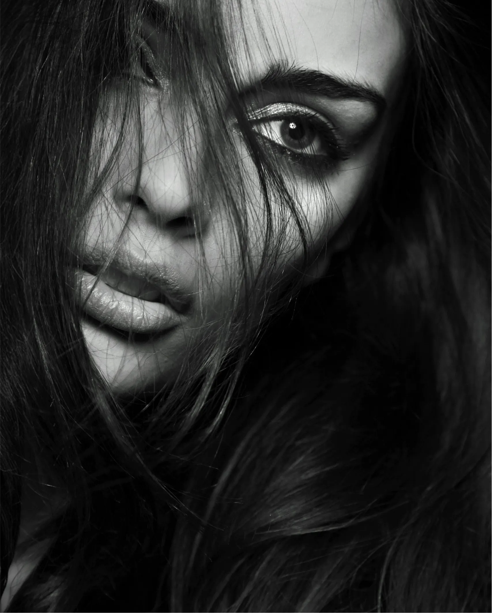 Black and white close-up of a woman's face partly obscured by long hair, highlighting one eye and lips.