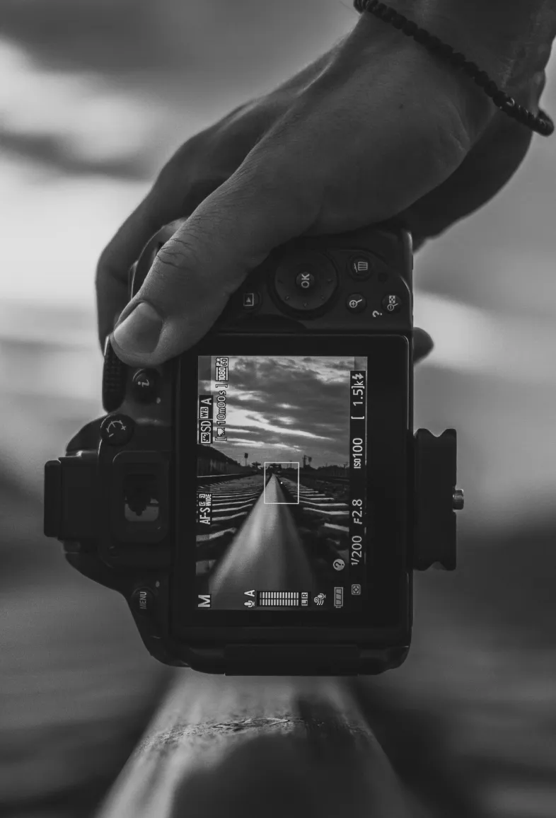Black and white photo showing a hand holding a digital camera capturing an image of railway tracks stretching into the distance under a cloudy sky, with the camera screen displaying the focused view of the tracks.