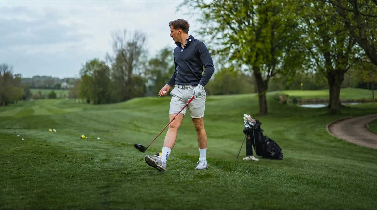 Man in black long-sleeve and shorts swinging golf club on green course with golf bag nearby.