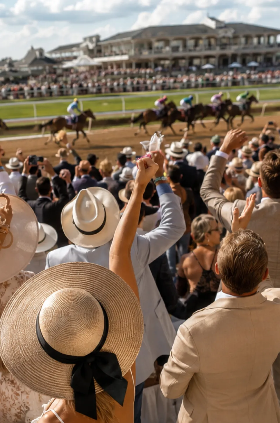 Crowd in hats and suits cheering at a horse race with jockeys riding on the track.